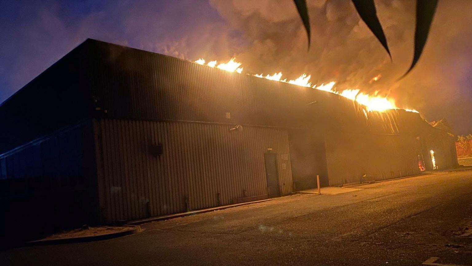 A warehouse on fire in Harwich. The fire service responded to the incident in the early hours of the morning therefore the picture has been taken in the dark, however, the warehouse is lit up by flames. A plume of smoke coming off the building is stretching into the sky. Where there are flames on the right hand side of the building in the picture, the roof has started to warp.