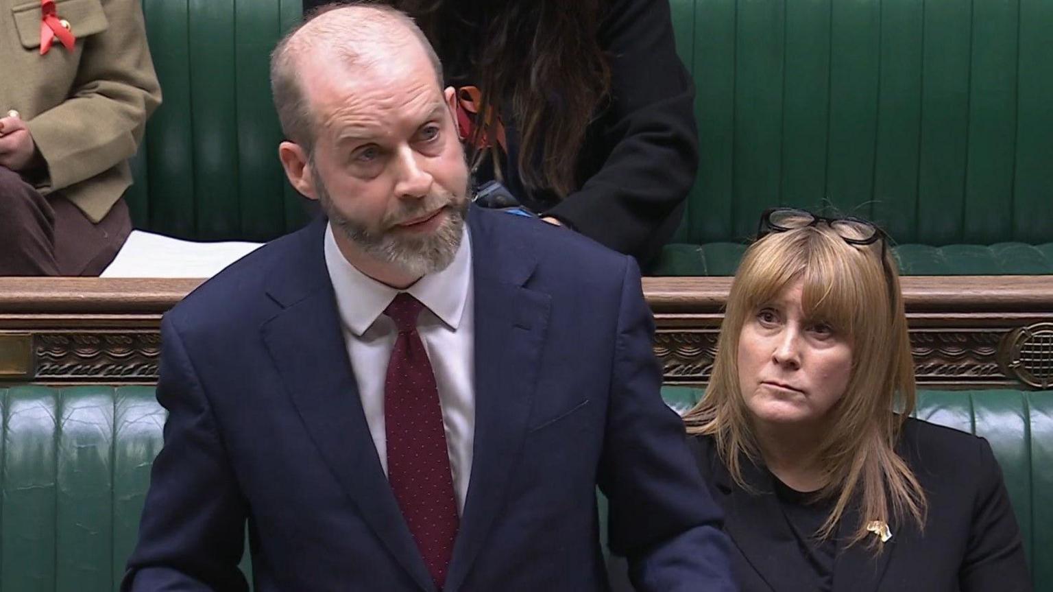 Jonathan Reynolds wearing a suit and tie standing in the House of Commons, with other MPs visible on the green benches