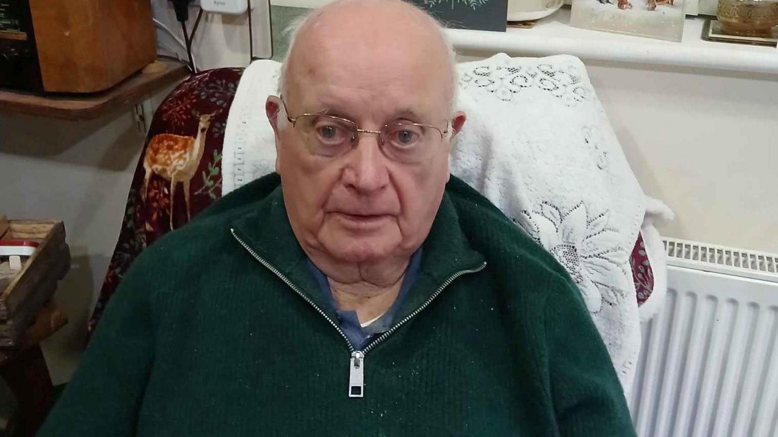 Michael Quirke is sitting in an armchair, smiling at the camera. He is wearing a dark green jumper with a blue t-shirt underneath and has reading glasses on his face.