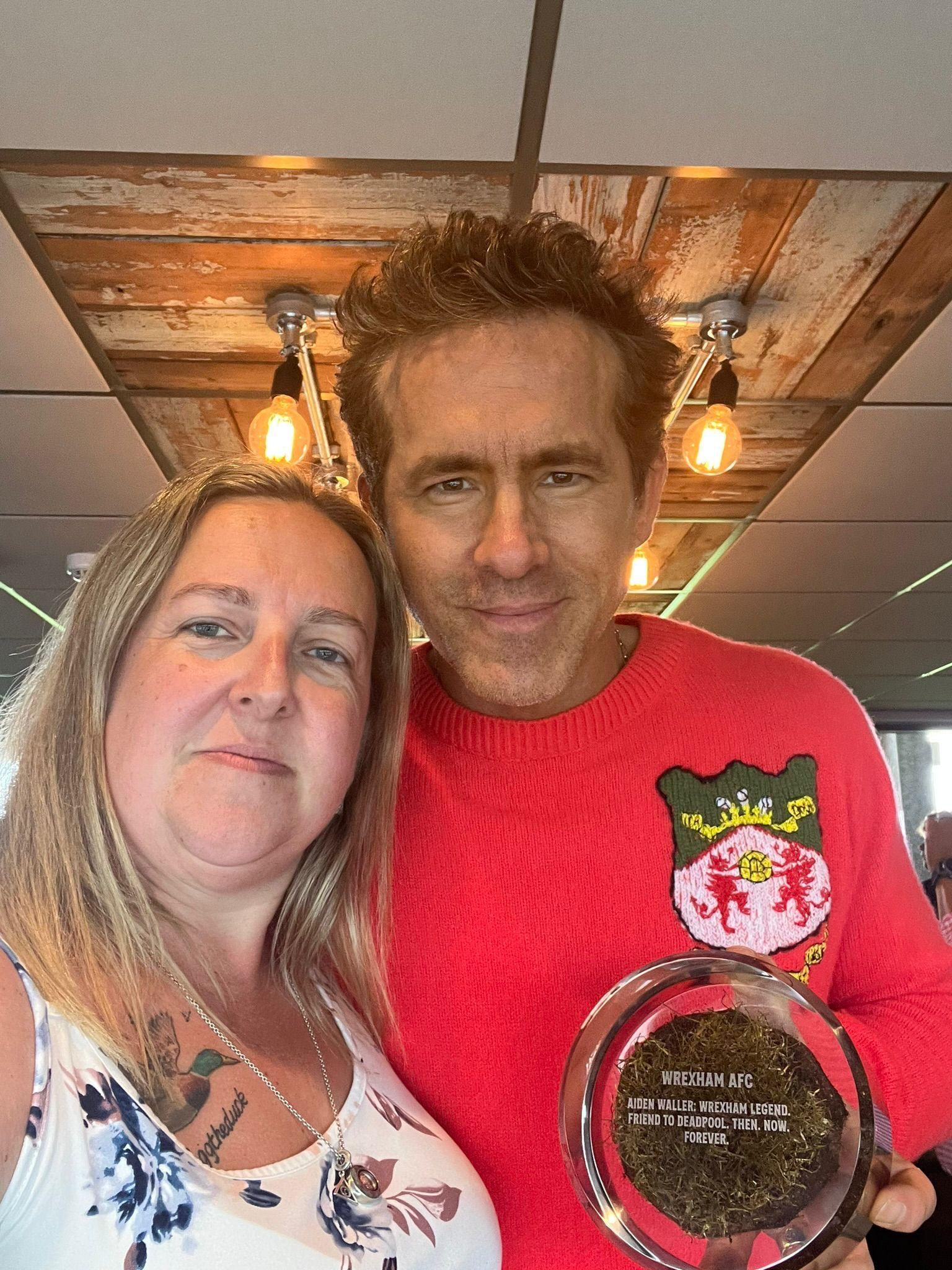 Sarah Waller pictured with Ryan Reynolds, who holds a circle glass plaque with a piece of turf inside and with writing inscribed on top. Reynolds wears a red knit jumper with the Wrexham shield, has brown hair and brown eyes and smiles at the camera. Sarah has blonde mid length hair and wears a white and blue floral vest top and looks at the camera. 