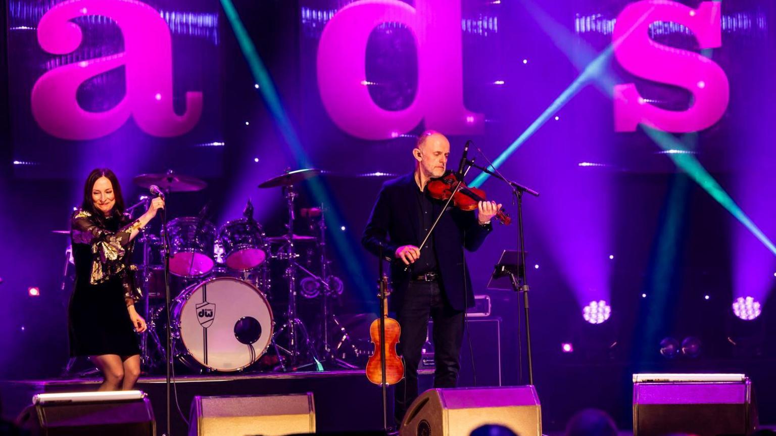 Fowlis and Chisholm are on a stage. There is a drum kit behind them and suspended from the roof as the letters "a", "d" and "s" from the word "Trads".