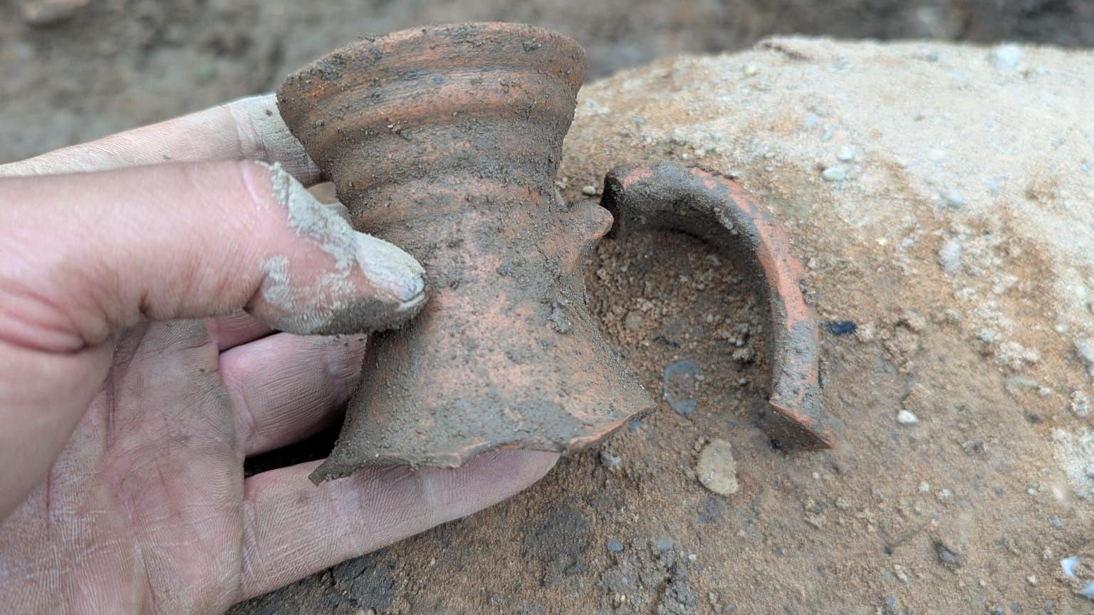A hand is holding a broken neck of a pottery flagon, with the remainder of it seen still partially buried in a trench.