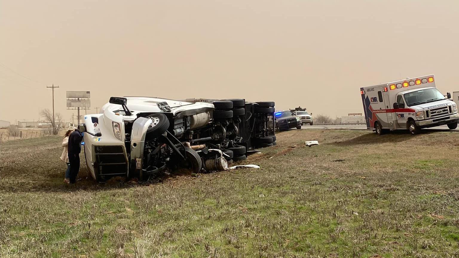 People inspect an upturned lorry on the side of a highway, with police and ambulance nearby.