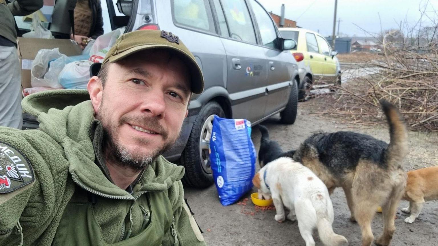 A man wearing a green hoodie and a green cap takes a photograph in front of some dogs eating food next to a car on a roadside.