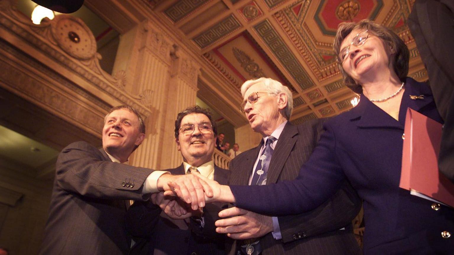 The SDLP team celebrating after ministerial positions were agreed on 29 November 1999.  Sean Farren is shaking hands with Bríd Rodgers. They are standing at either side of John Hume and Seamus Mallon in the Great Hall in Stormont. 