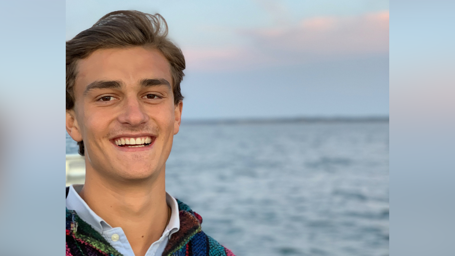 Henry Keith smiling. He has brown hair and is wearing a blue shirt with a colourful jumper on top. Behind him is what appears to be the sea