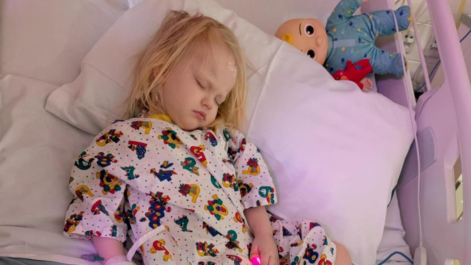 Molly asleep in a hospital bed wearing a gown. She has shoulder-length blonde hair and where's a colourful hospital gown. A doll lies near her head beside the pillow.