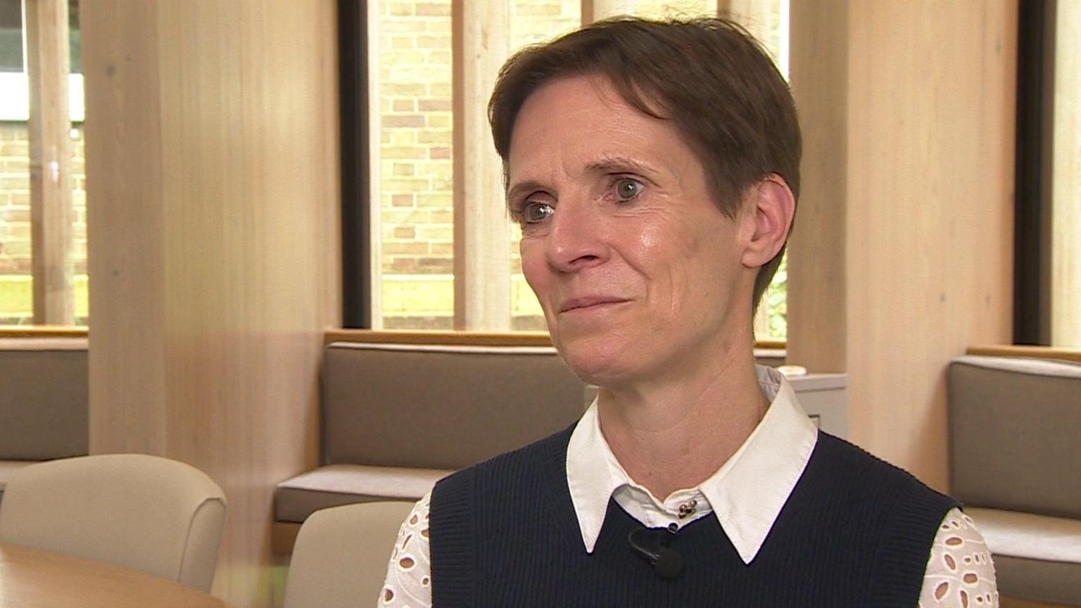 Head teacher Jane Gandee sitting in front of a window. She has short brown hair and is earing a dark coloured top with white collars