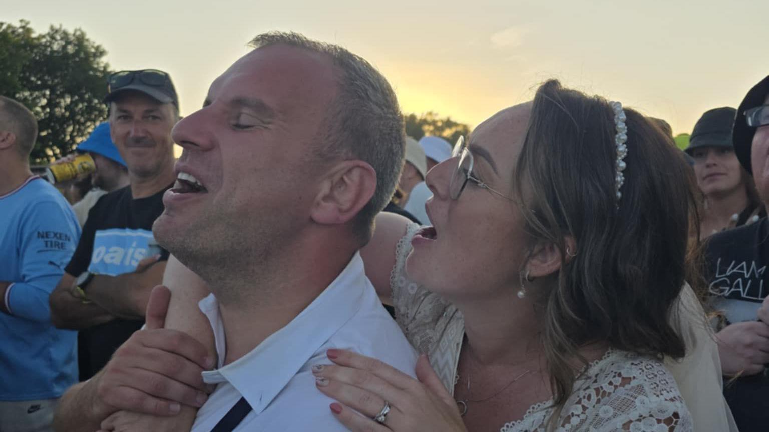 Gareth and Kim Gladman singing along to the Oasis performance in the crowd. She has her arms clasped around him, with other fans around.