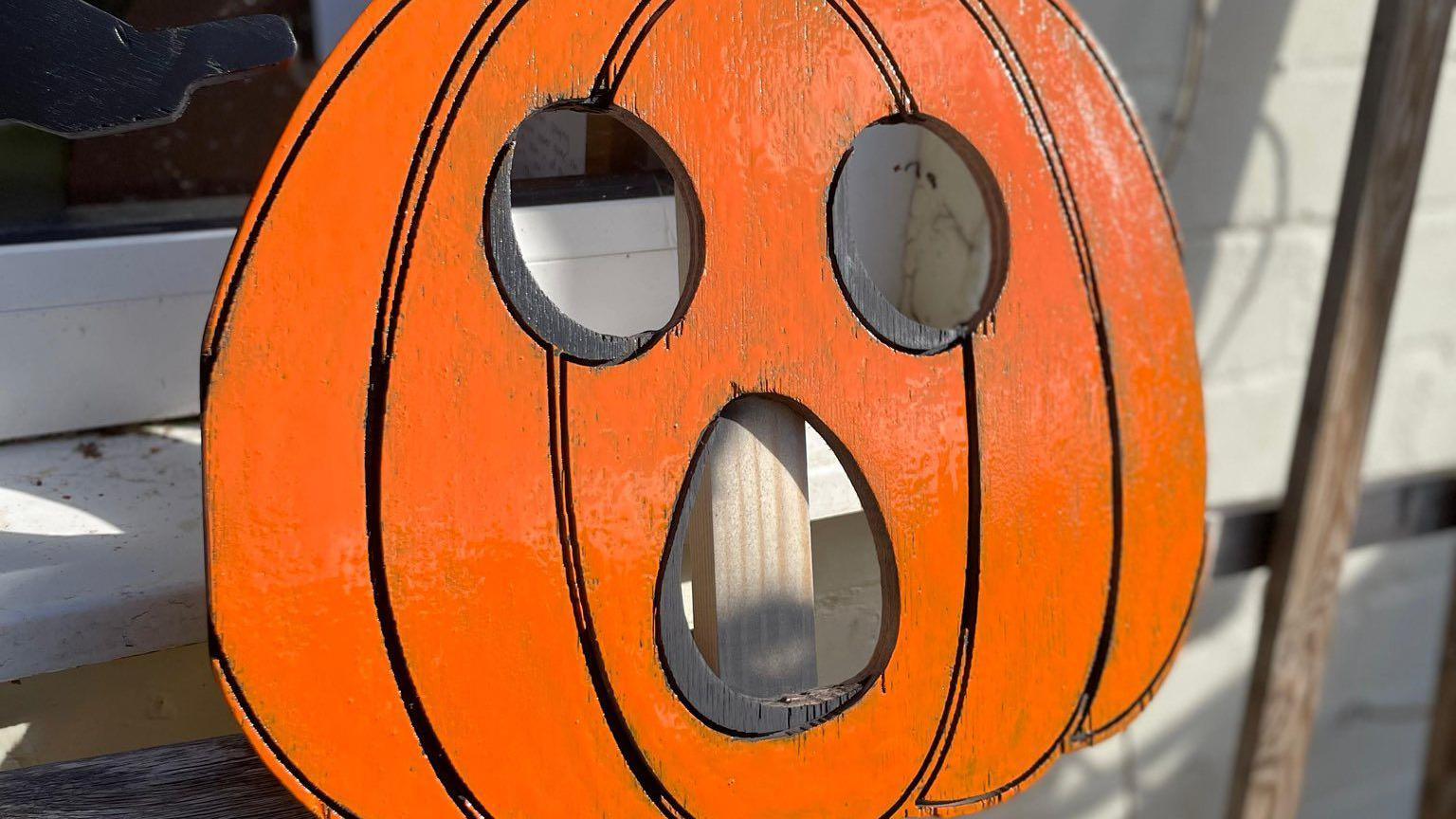 A flat piece of wood cut in the shape of a pumpkin with a face carved into it. It is painted orange