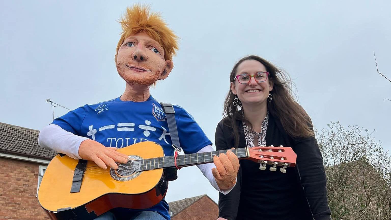 A woman - Sharon Kulesa - smiling at the camera while standing next to an effigy of Ed Sheeran which is wearing an Ipswich Town top and holding a guitar.