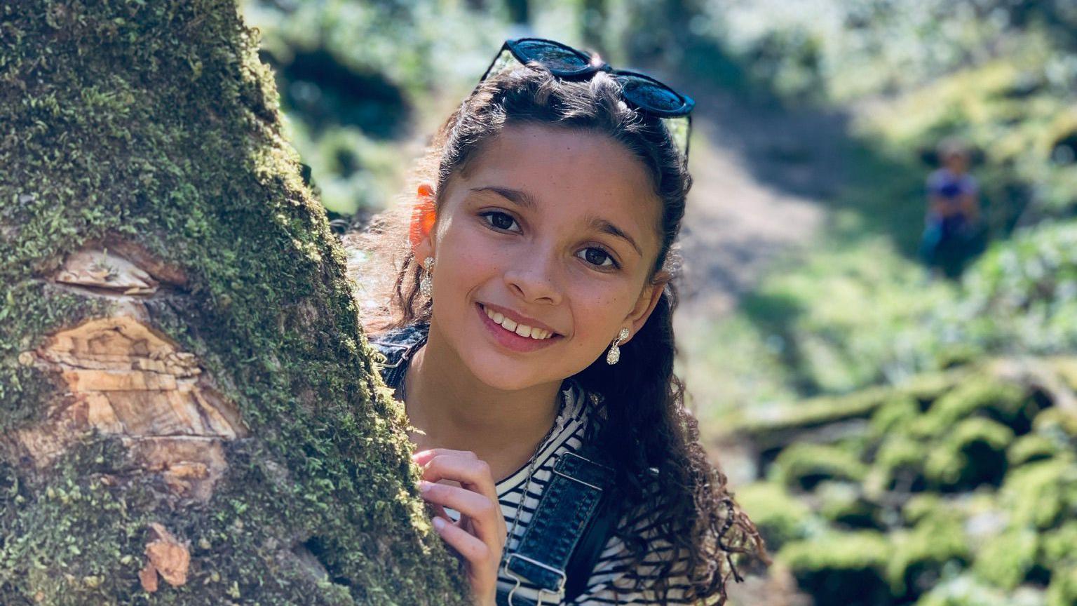 Amelie peeking out from behind a tree in the woods and smiling at the camera. It is a picture taken before her diagnosis. She has long and curly dark hair which is pulled back by the sunglasses propped on top of her head. She has sparkly dangling earrings and is wearing a striped top with denim dungarees. 