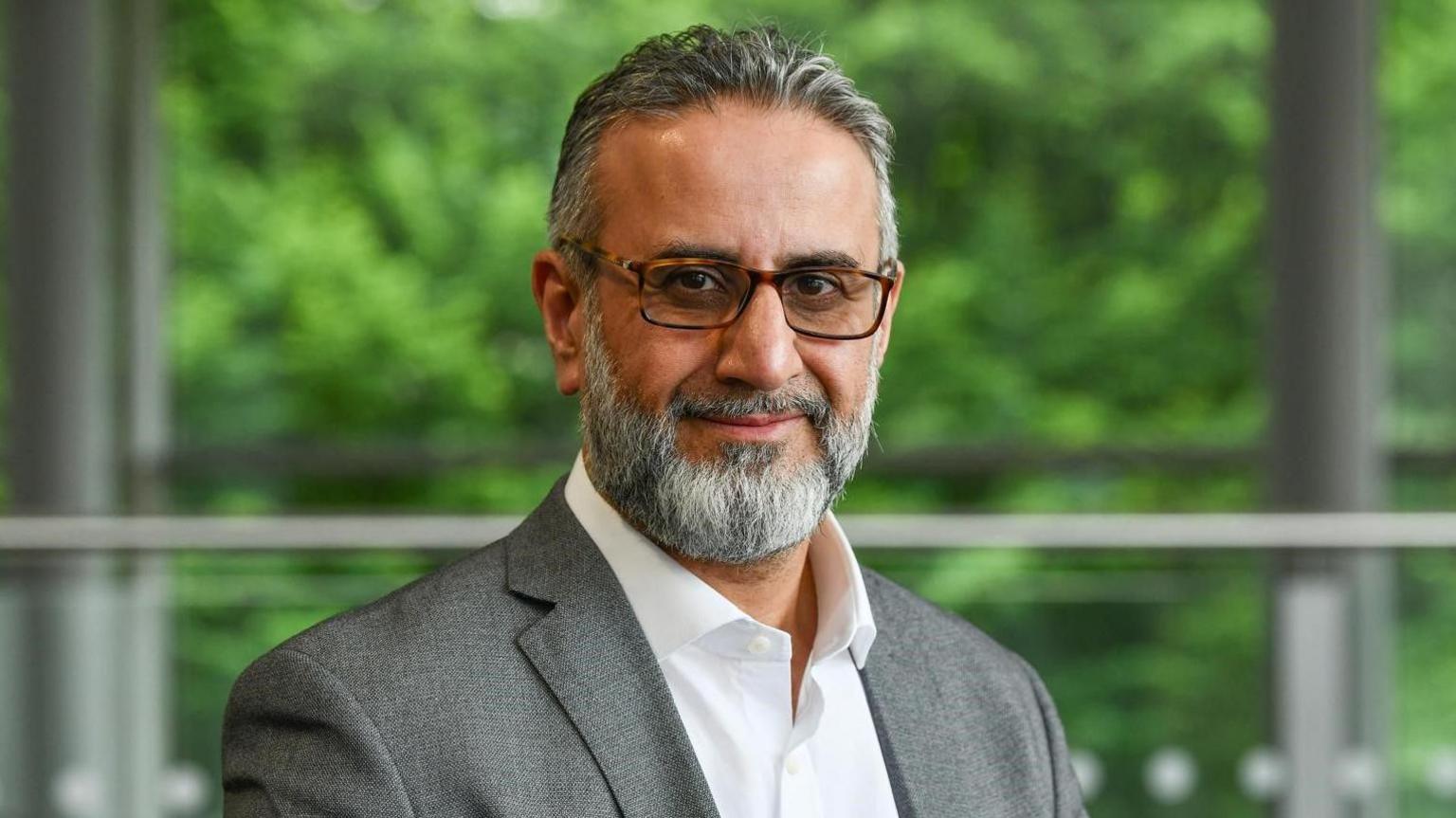 Dr Belal is standing in an office. He is wearing a grey suit and a white shirt. He has short grey hair, a grey beard, and is wearing tortoiseshell glasses. Trees are visible through the window behind him
