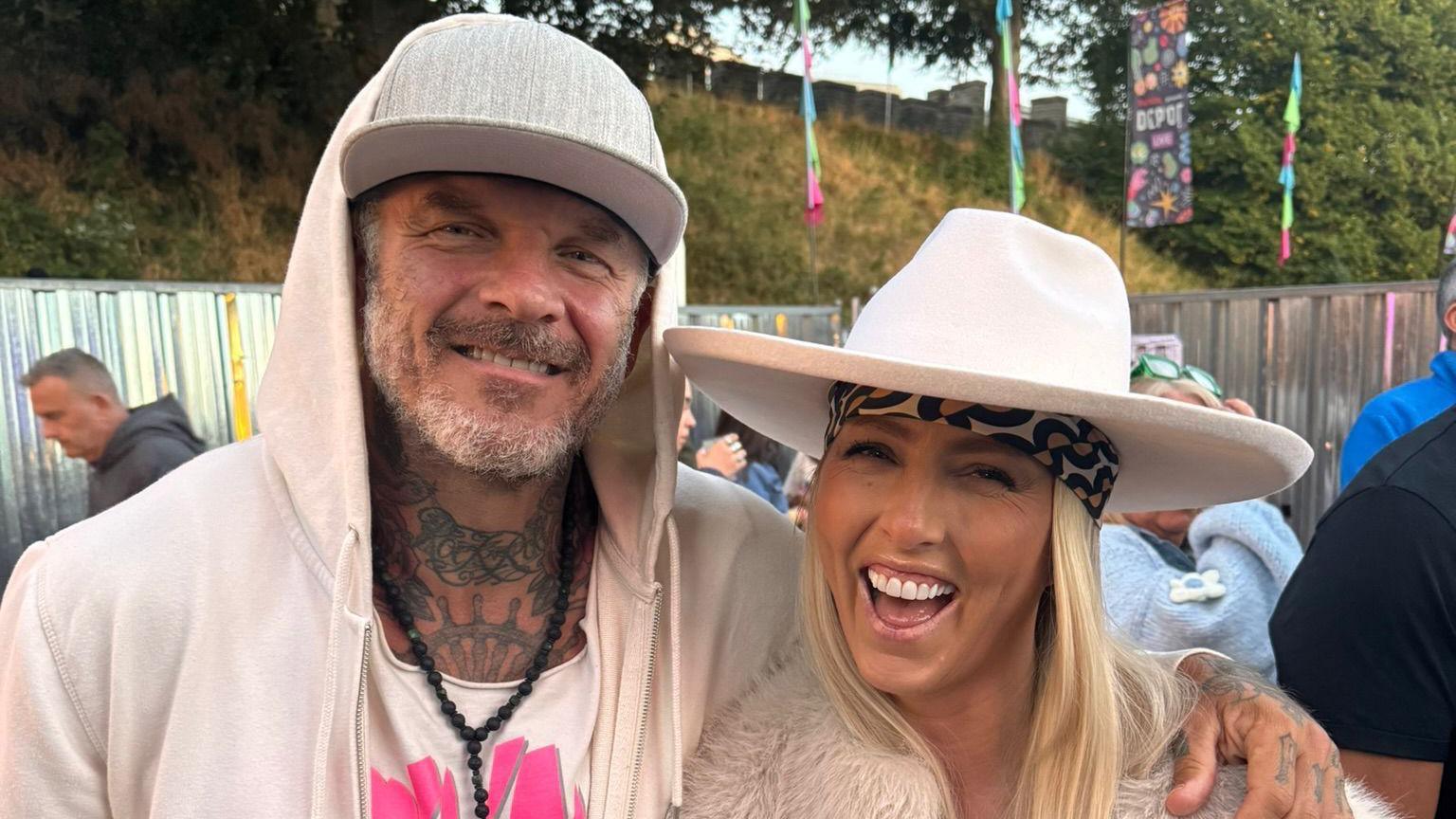 Photograph of Mathew Pritchard and his partner Ciara Dunne. Mathew wears a cream zip up hoodie, a white vest shirt and a black beaded necklace. He wears a grey flat cap hat, has a silver beard and blue eyes and smiles at the camera. His wife wears a patterned scarf under a white wide brimmed hat, and a fur jacket.