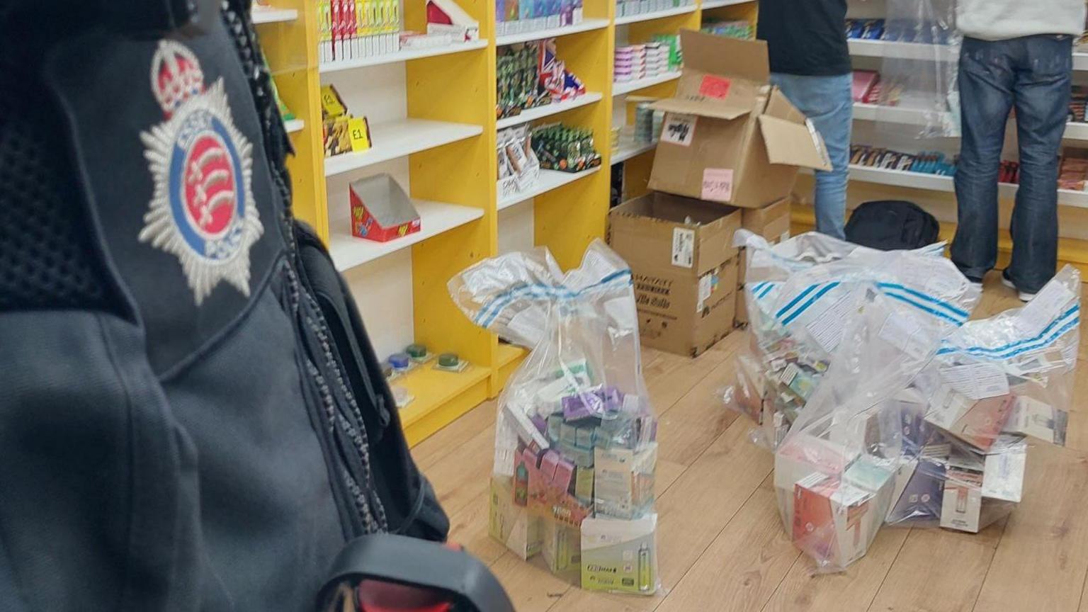 A police officer, to the left, and only showing his body, in uniform, in a shop, with two other people. There are items on the shelves, and lots of items in bags on the floor and three cardboard boxes. You can only see the legs of the two people.