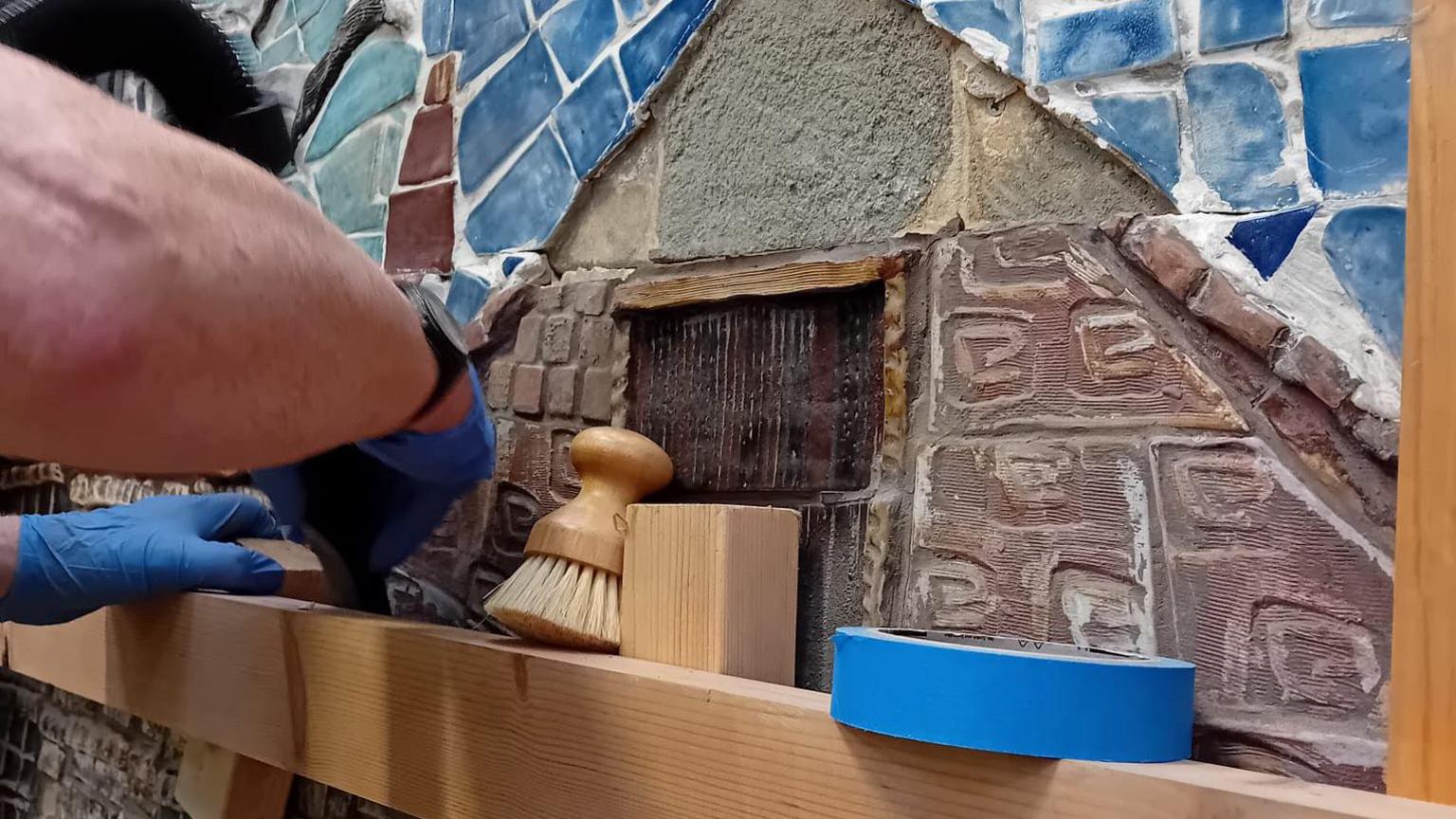 A woman with blue gloves using a wooden frame to piece back together a colourful mural