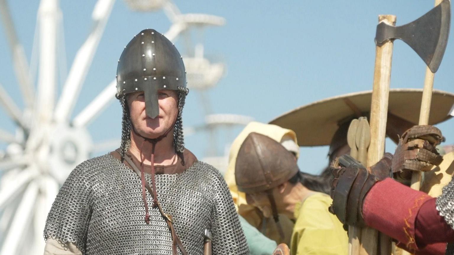 Man in chainmail tunic dressed like a Viking with a metal helmet stares at the camera. He is surrounded by others dressed in Viking-era costume. Someone is holding a large axe. 