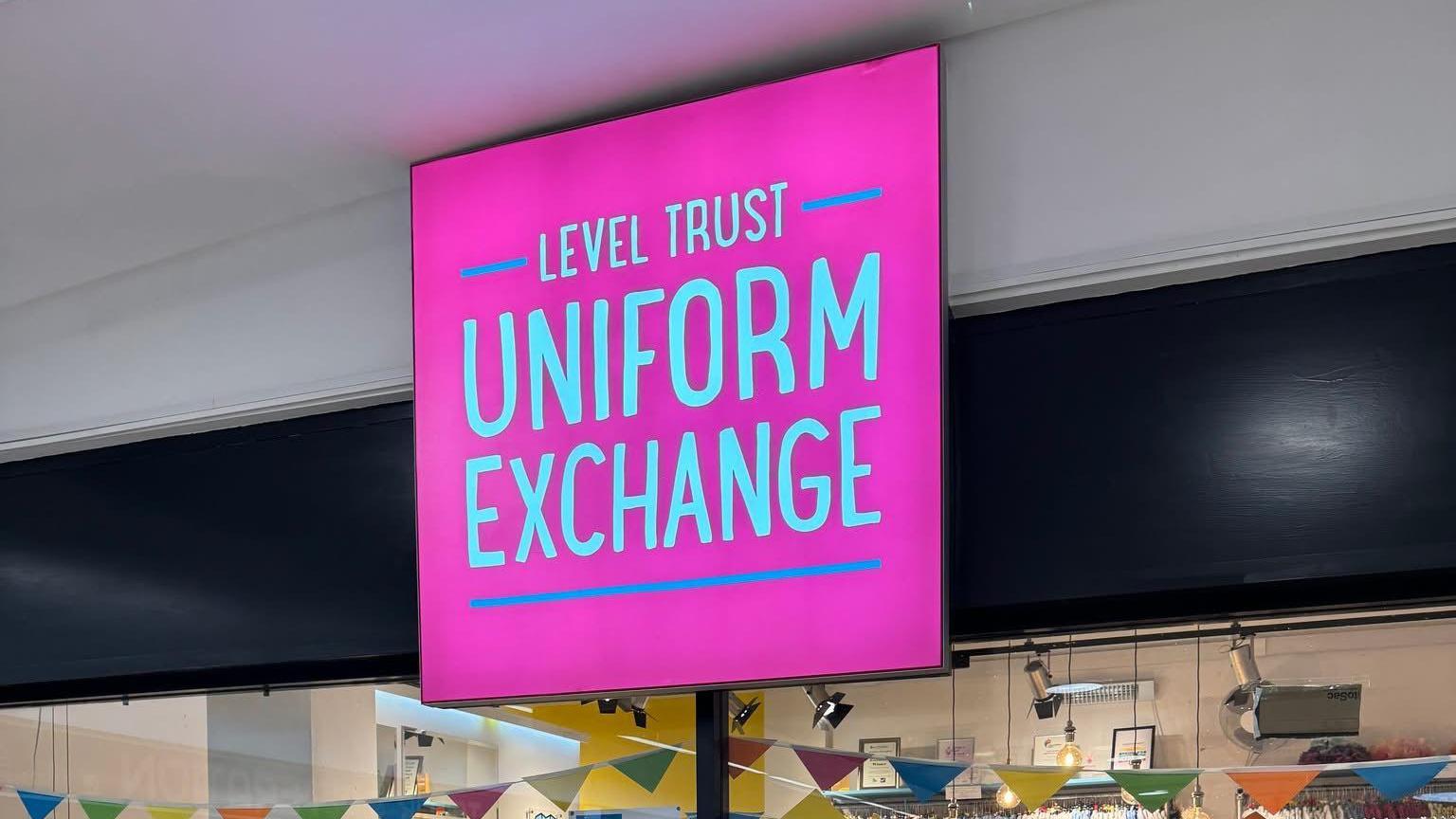 A pink sign that reads "Level Trust Uniform Exchange" which is hanging above a glass-panelled shop front. Bunting can be seen inside the shop, hung from the roof.  