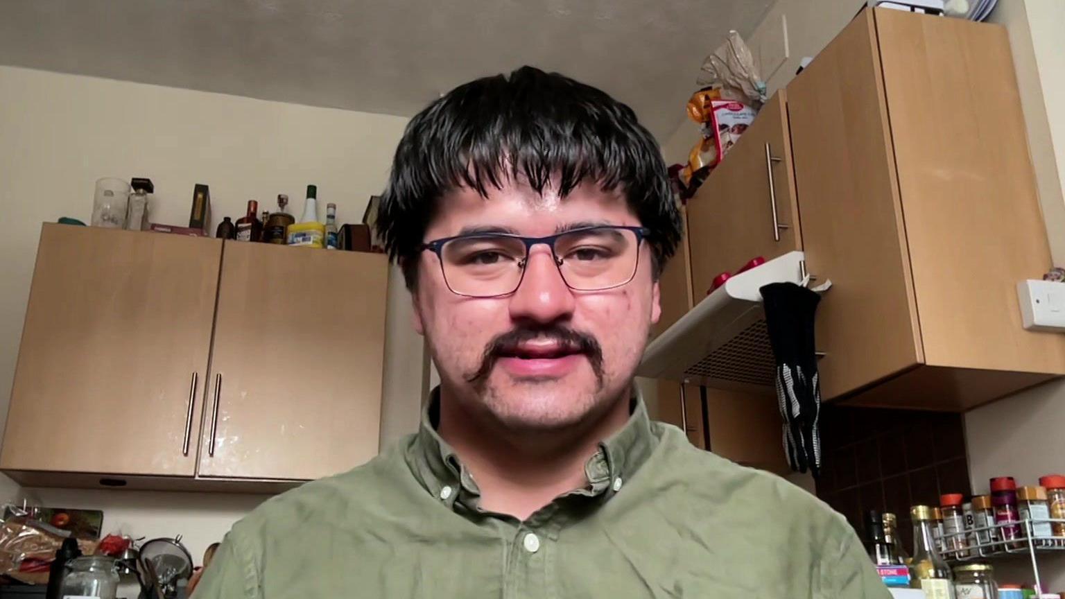 A head and shoulders photo of a man sitting in a kitchen. He has black hair and a black moustache. He is wearing glasses and a green shirt. Wooden kitchen cupboards, bottles, spices and tinned food are behind him.
