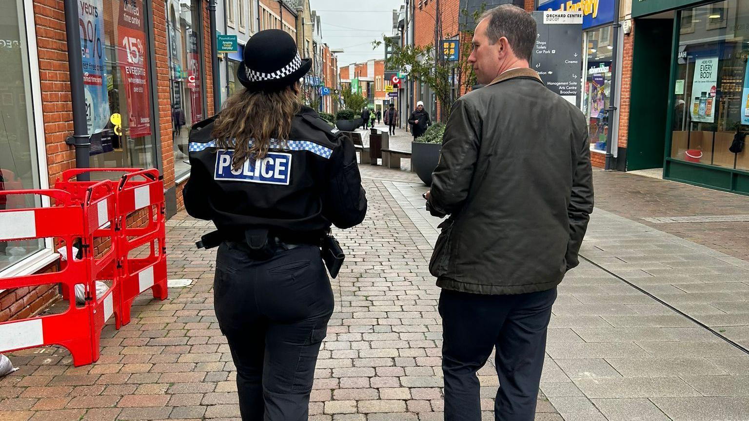 PCC Matthew Barber with female police officer in Didcot. They are photographed from the back. They are passing by shops. It is a cloudy day.