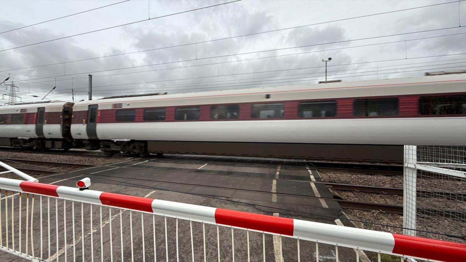 A red and white liveried trains speeds over a level crossing, with the red and white road barrier in the forefront of the photo.