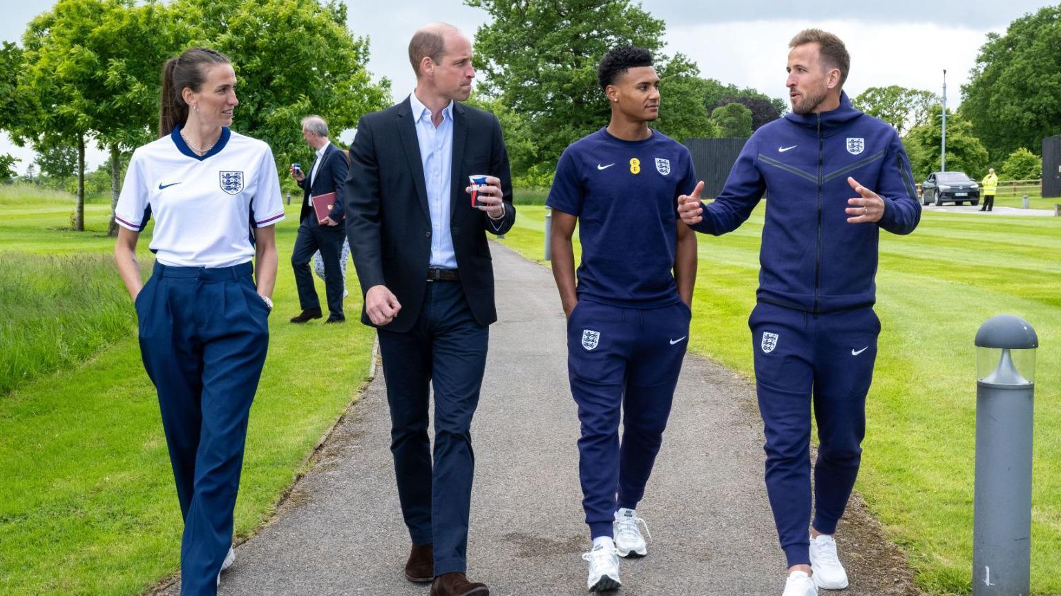 Three men and a woman walk down a concrete path in a grassy landscaped area. 