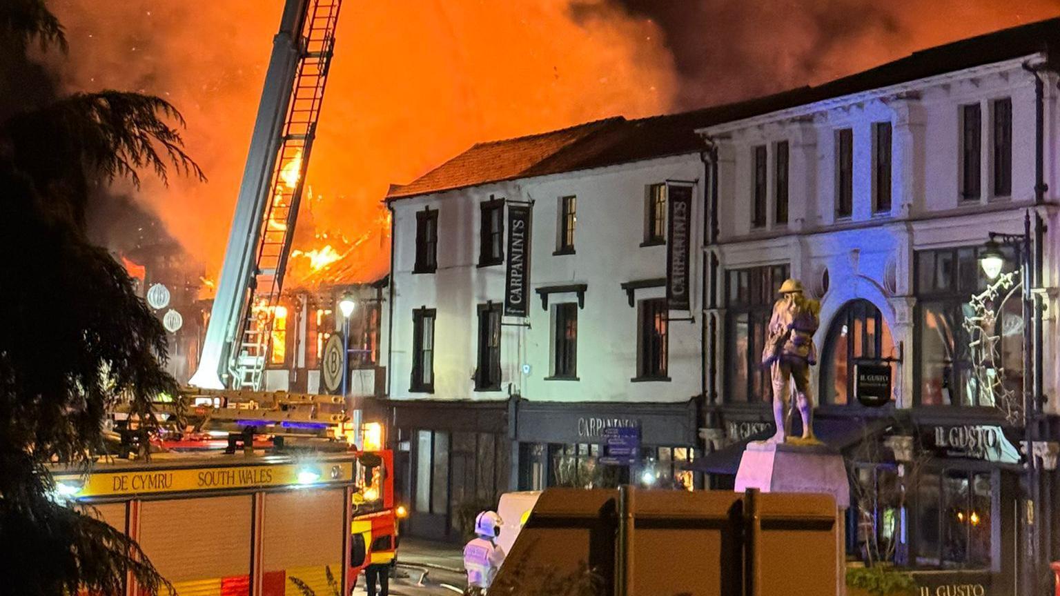 Fire trucks and fire crews in Abergavenny town centre with large orange flames and smoke in background