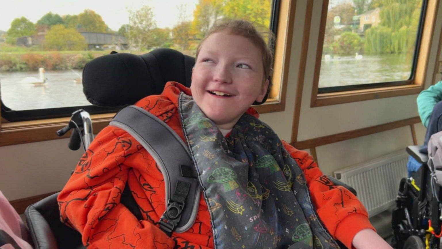 A picture of Declan sitting in his wheelchair on the boat wearing a red top and smiling.