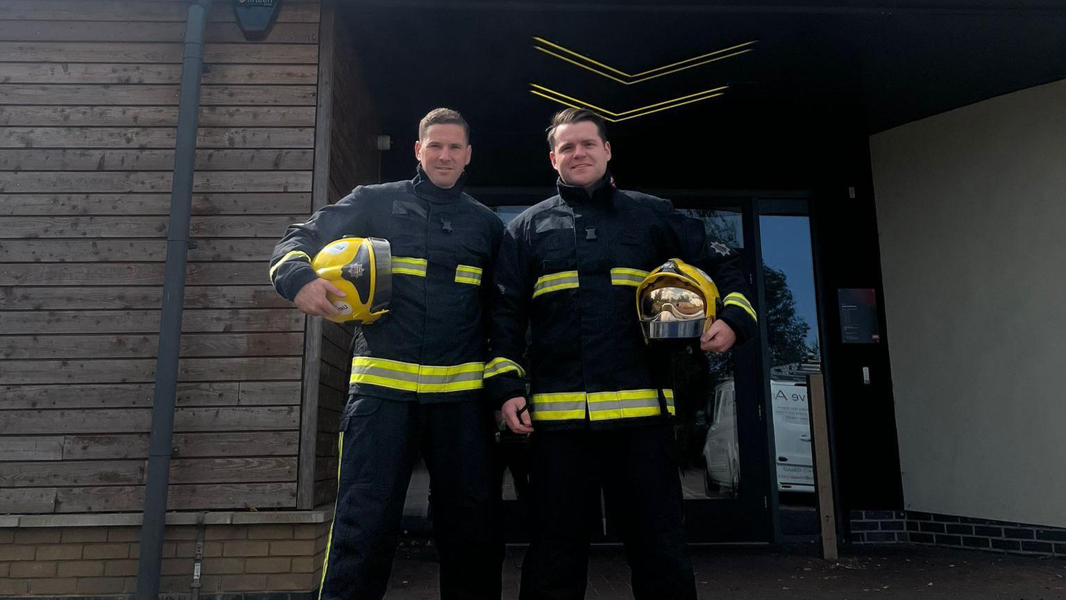 Jonathan Freeman and Sam Hayward standing outside a gym holding their helmets and wearing fire gear