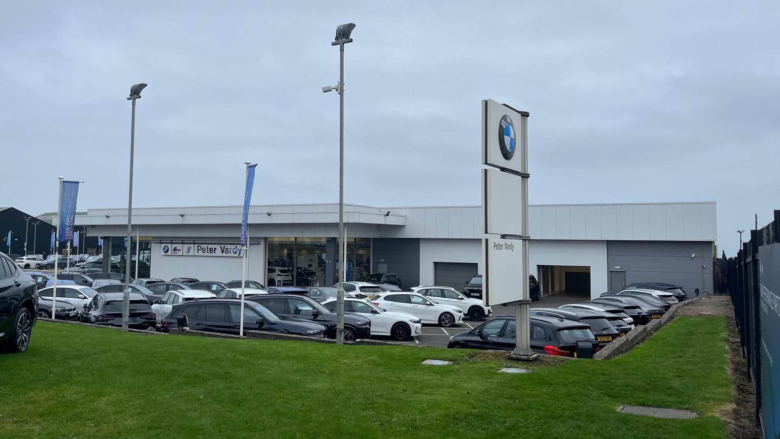 Lots of parked cars outside the Peter Vardy BMW showroom. There is BMW sign in the foreground and blue banners flying.