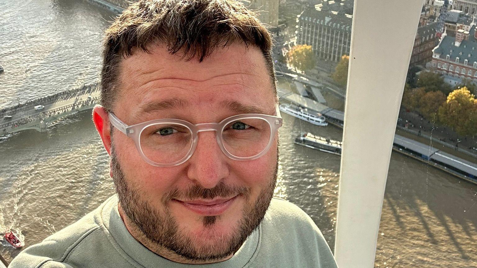 This is a selfie of Mark Lewis, taken against the backdrop of Westminster, in London. He's a white man, in his 40s, and wears a grey sweatshirt with transparent round glasses. He has dark hair and dark facial hair too.