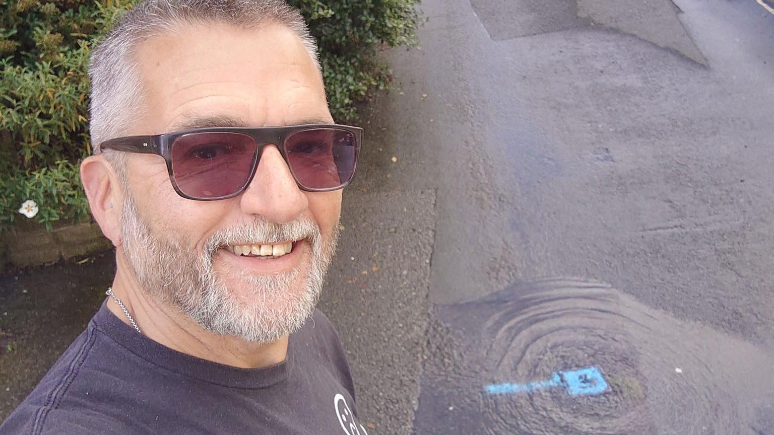 A man with grey hair and a beard, wearing sunglasses, takes a selfie next to a flooded area of road with the leak point marked in blue. 