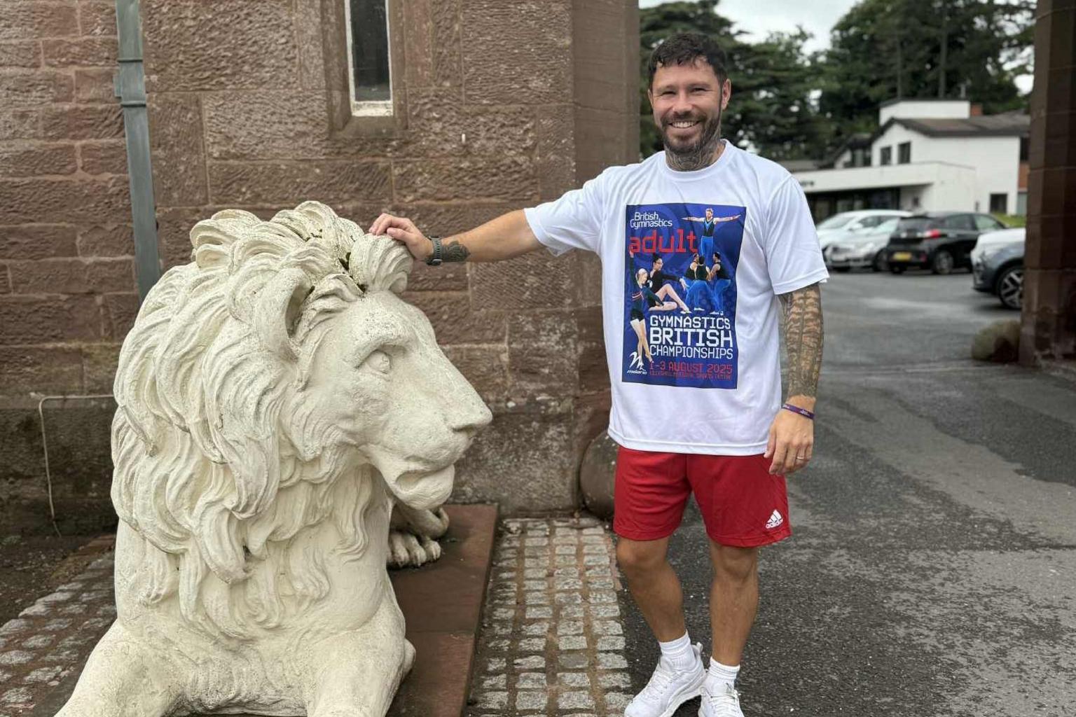 A man with a white T-shirt with a British Gymnastics Championships picture on it. He has short brown hair, a brown beard and tattoos on his arms. He has red shorts, white trainers and white socks. He is leaning on a white lion statue. He is outside on tarmac in front of a brick building. There are vehicles in the background.
