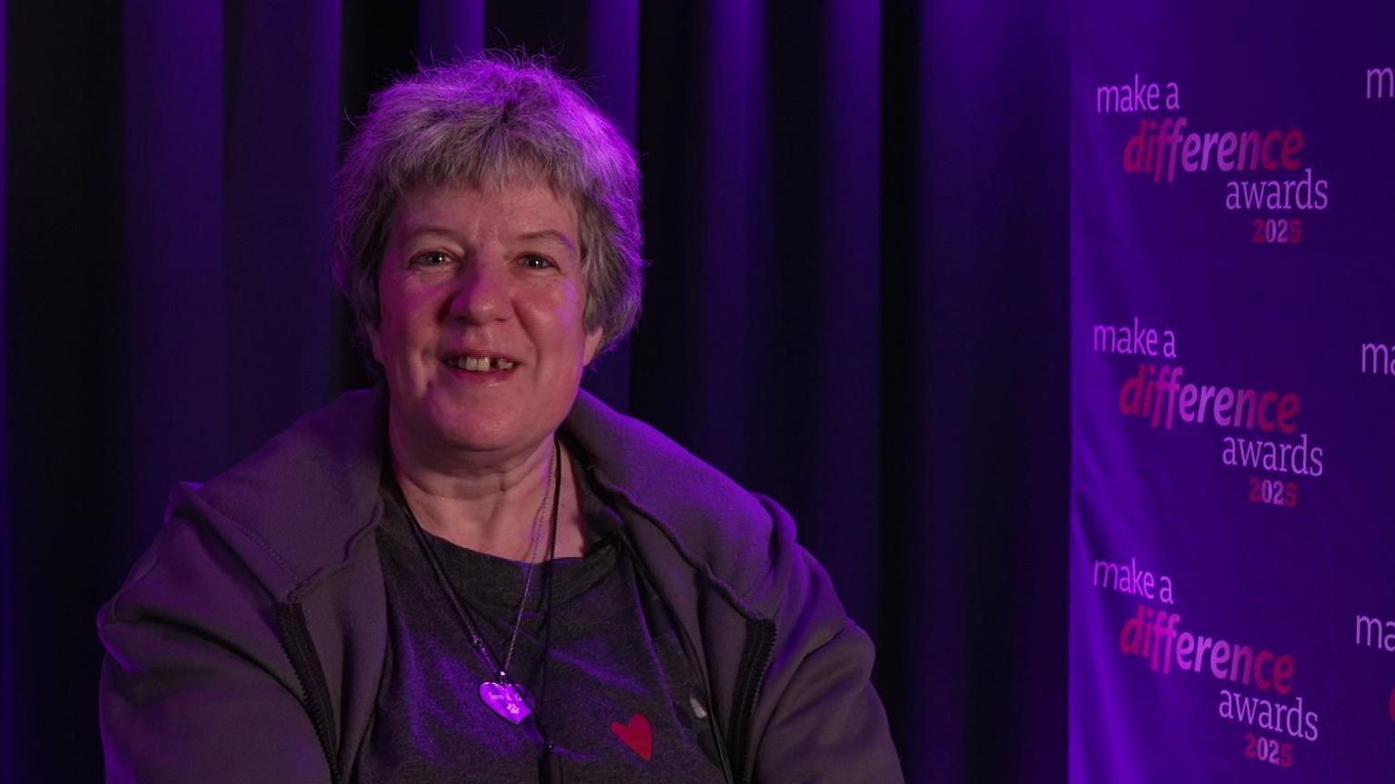 Women with short grey hair smiling at camera. She is wearing a heart neckless and a grey jacket. She is sitting down and is in front of a black curtain with the make a difference awards poster to her left shoulder.