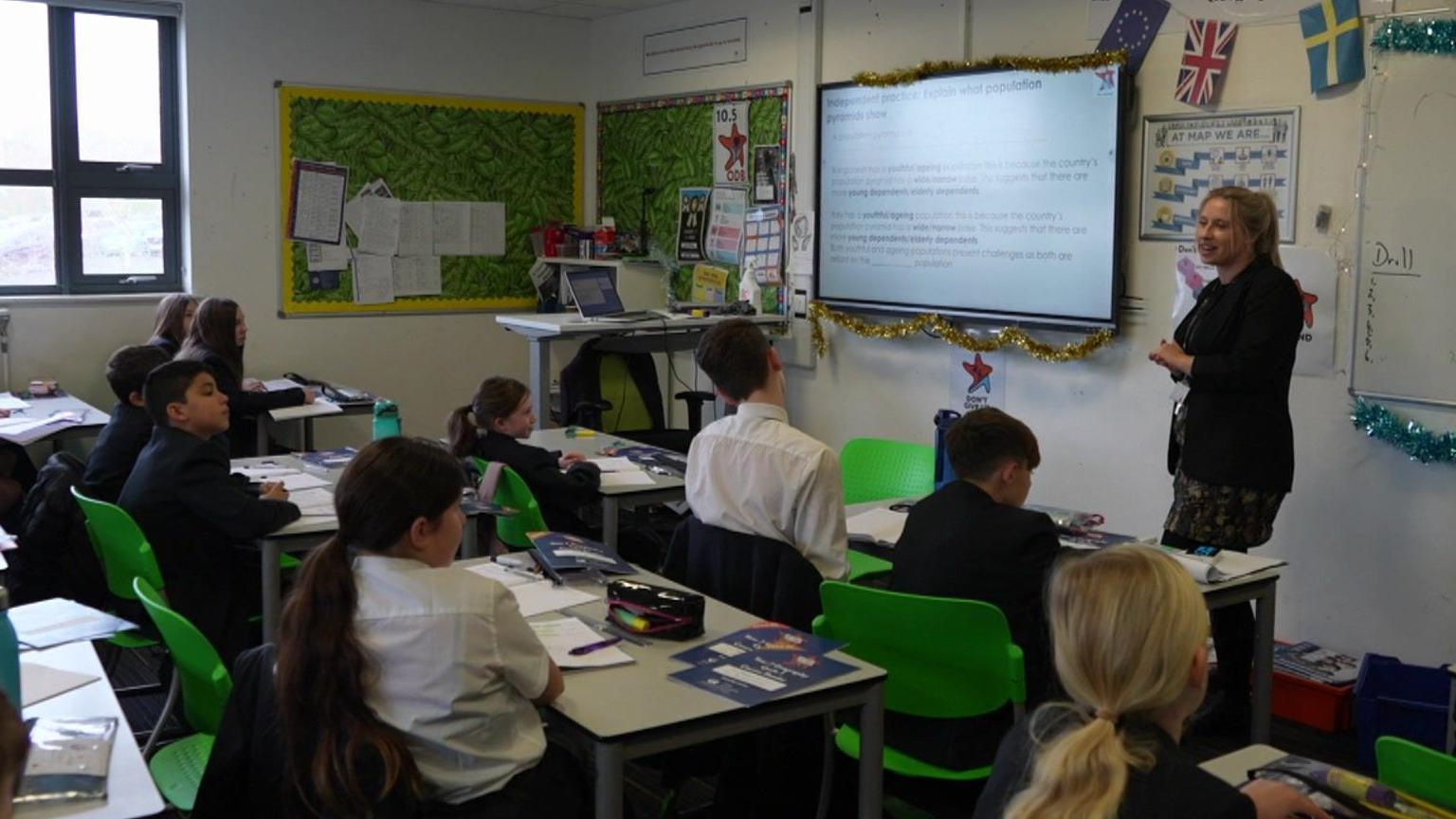 A teacher wearing a grey dress and a black blazer stands at the front of a classroom while pupils listen to her while she talks during a lesson with information on the interactive whiteboard to her right.