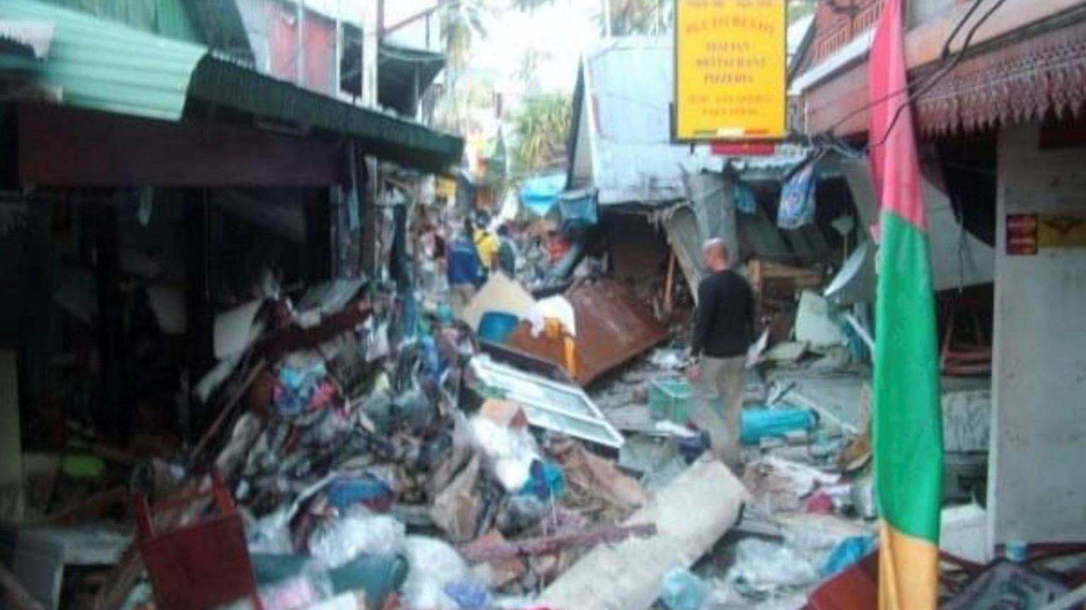 A narrow street in Koh Phi Phi market that has been completely destroyed by the tsunami. A man wearing a black long sleeved shirt is standing amongst the debris. The road is littered with chairs, broken furniture and rubbish.