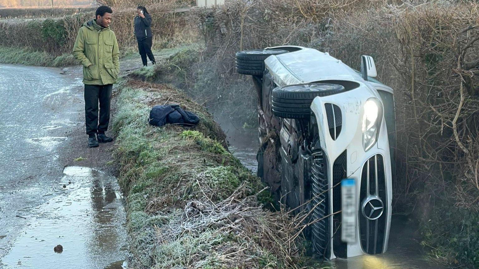 A white Mercedes car overturned on its side in a river stream. A man is seen on the edge of the road staring at the car.