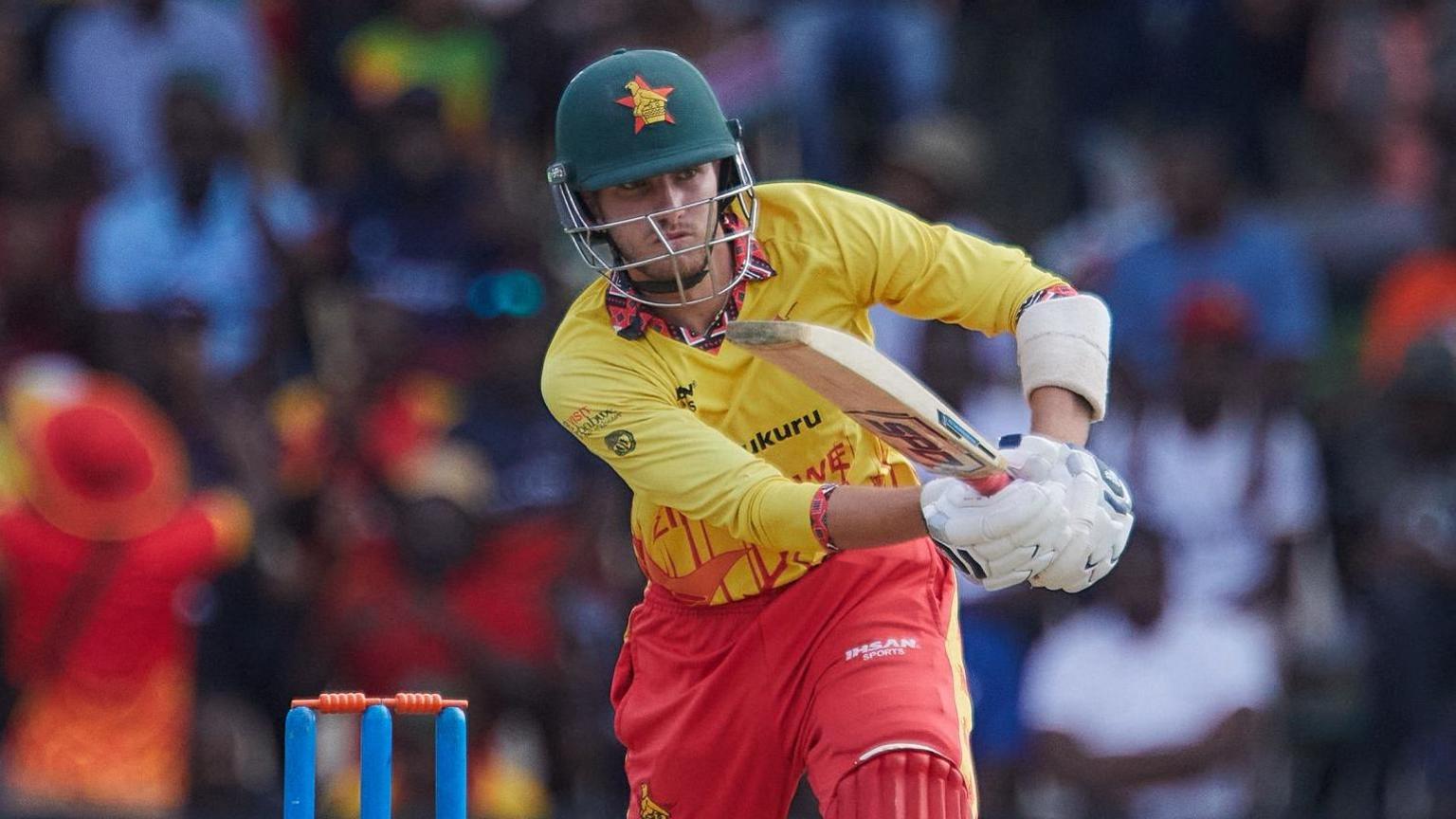 Brian Bennett, wearing a green cricket helmet with a Zimbabwe crest on the front, a yellow Zimbabwe shirt, red trousers and red pads, is seen holding a bat while playing a cricket shot on the front foot. Blue stumps with orange bails are seen behind him, with a crowd out of focus in the distance