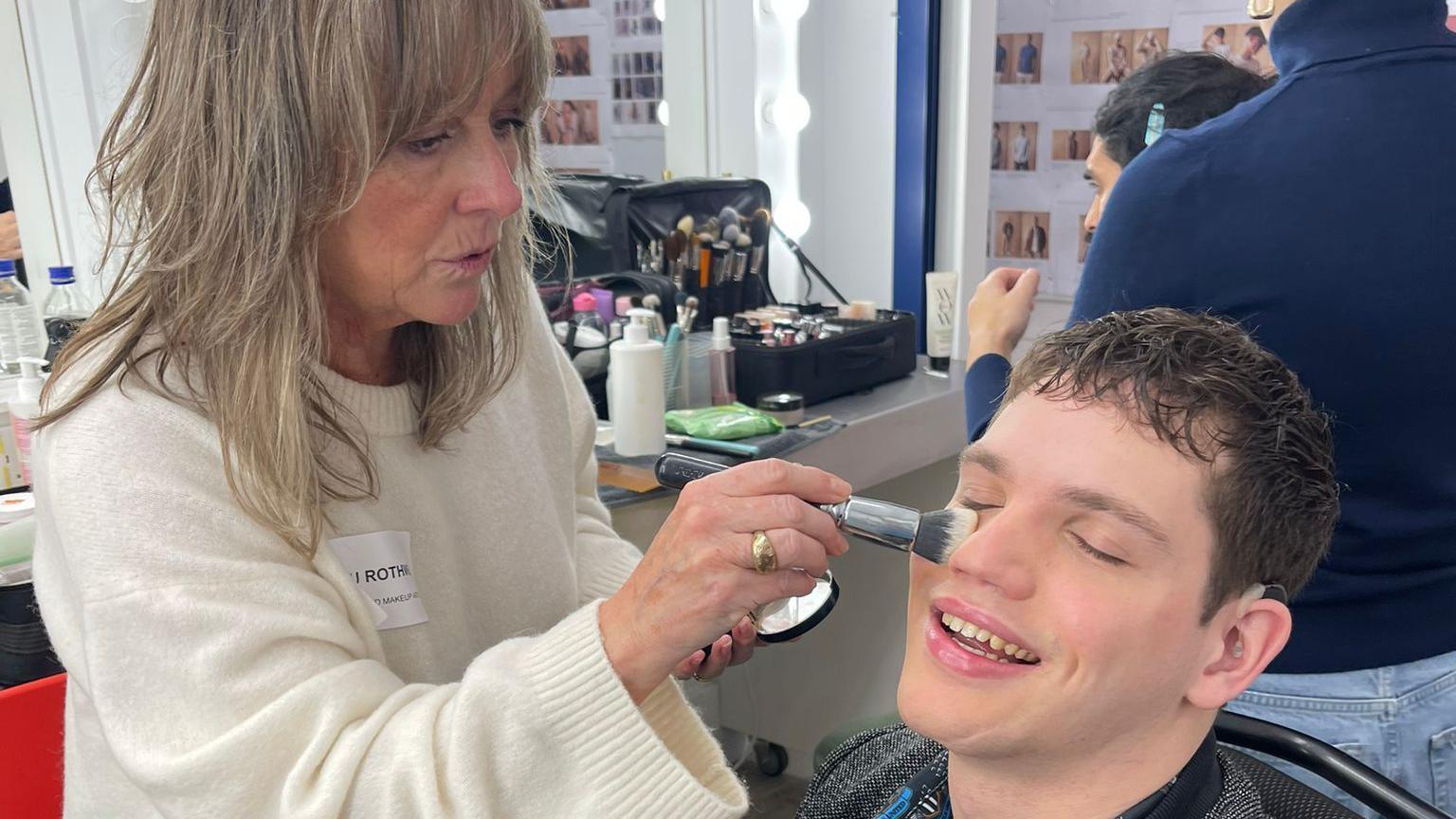 A man, Elliot Caswell, is sitting down, having his makeup done in a dressing room by a makeup artist. He is wearing a grey jacket and a patterned shirt. He is smiling as the makeup artist applies powder to his face.