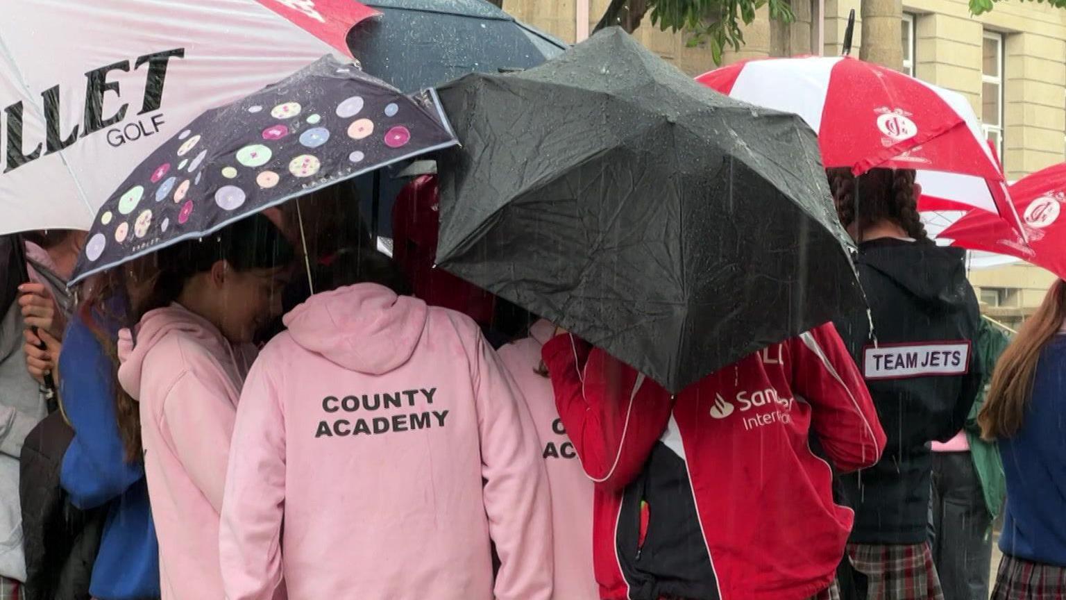 People in pink, red and black netball hoodies standing under umbrellas in the rain.