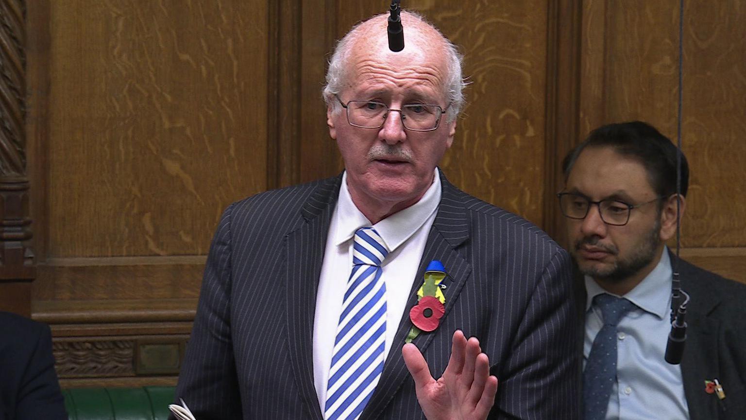 A man with white hair and glasses looks across the screen. He is wearing a navy suit with a white stripe and a blue and white striped tie. He is wearing a poppy and a pin with Ukrainian flag colours. 