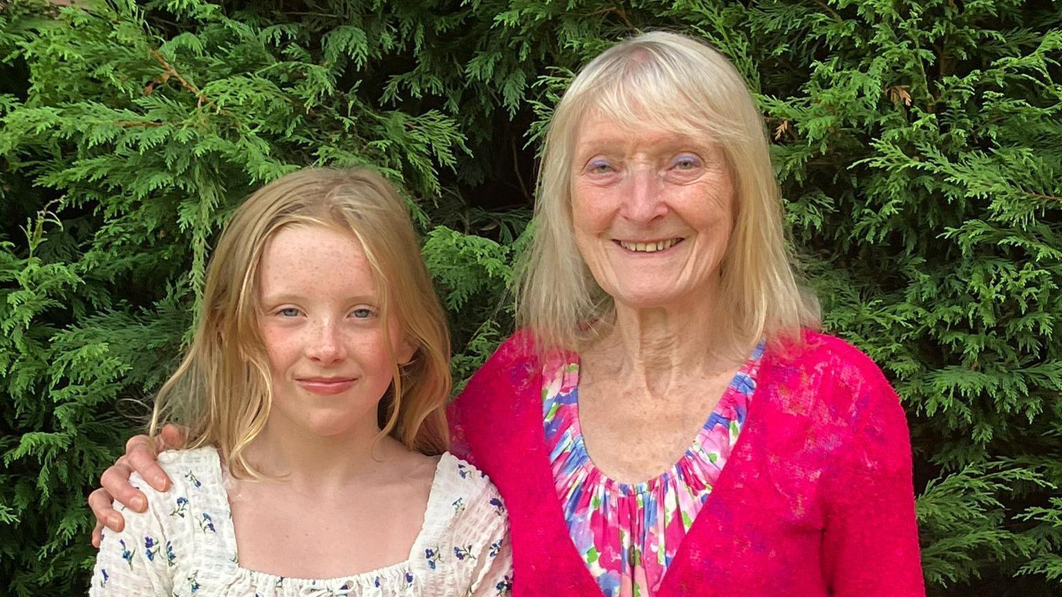 Pauline wearing a pink blouse with her hand over granddaughter Connie's shoulder as the two pose for a photo in a garden with a green hedge seen behind them.