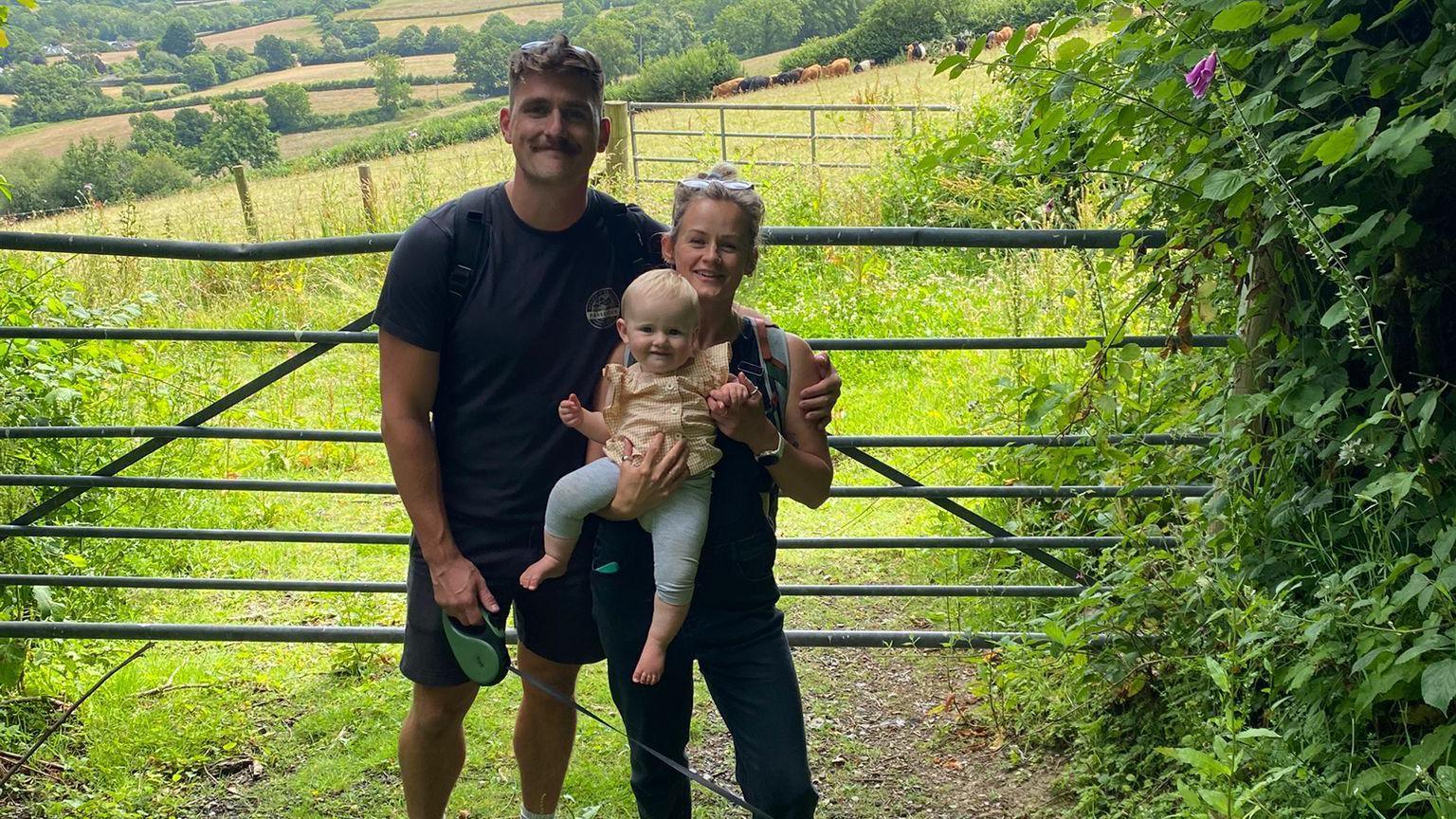 A man and a woman holding a baby standing in front of a field