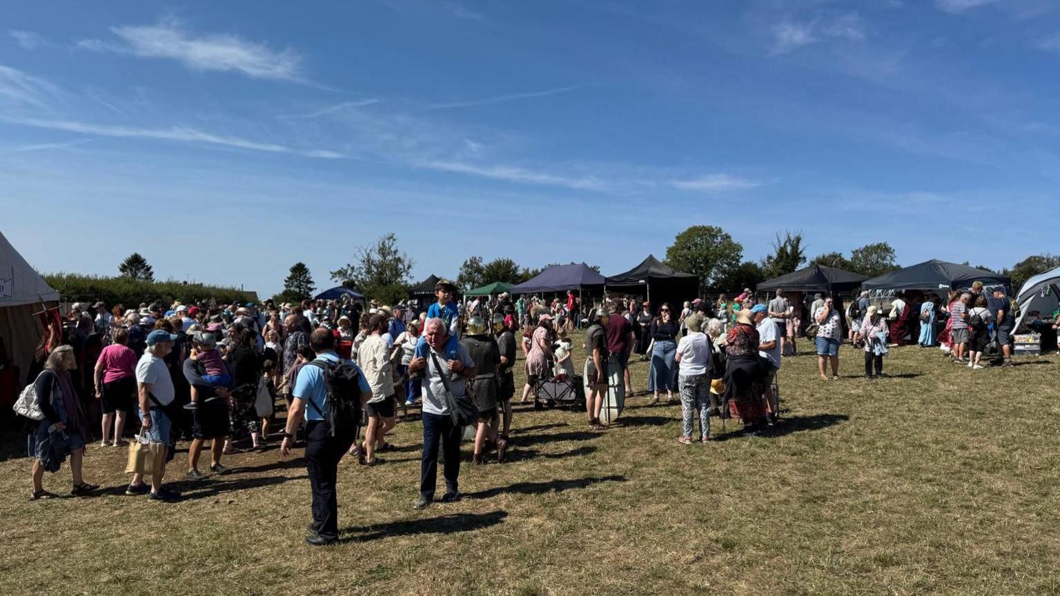 A large crowd of people move around a field on a sunny day. There are tents set up along one side of the field, as you'd expect to see at a festival. 