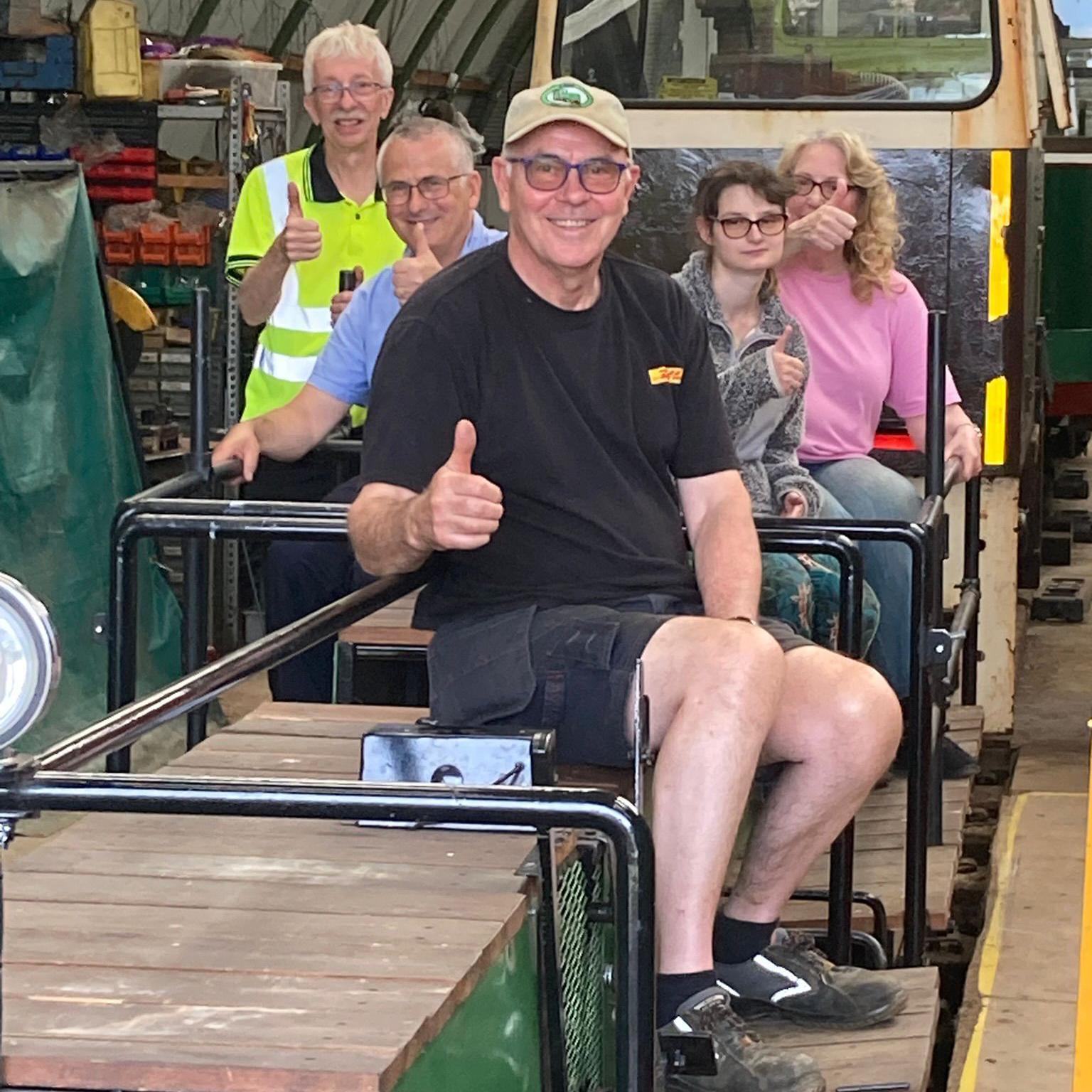 Three men and two women sit on a railway wagon giving the thumbs up. They are all wearing casual clothes except one, who wears a yellow hi-vis vest. The wagon is part of a train with a cream metal and glass cab. They are inside an engine shed with a curved roof.