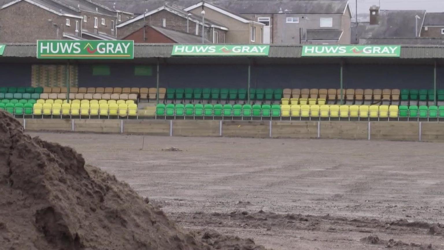 Eisteddle presennol yn safle CPD Caernarfon gyda stad dai yn y cefndir. Yn y blaen mae rhan o gae'r Oval sydd yng nghanol cael ei lefelu fel rhan o gynllun uwchraddio ac mae tomen o bridd yng nghornel chwith gwaelod y llun.