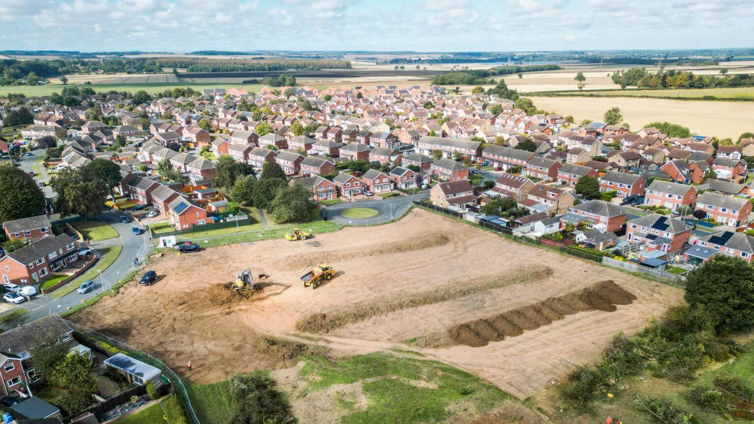 A drone view showing diggers and lorries moving around a site of freshly dug earth, which backs on to a housing estate, with fields and low hills in the background.