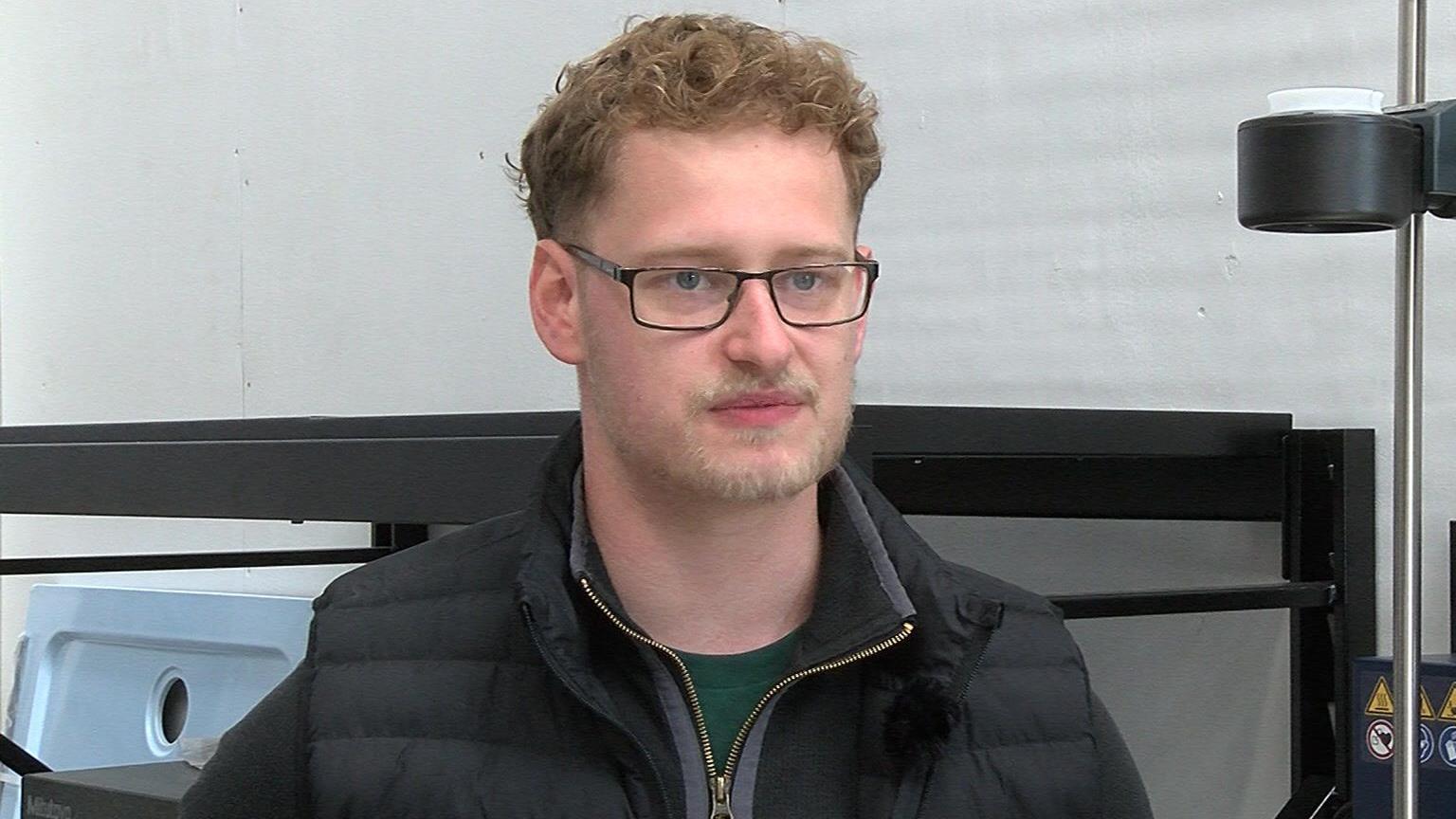 A man wearing a grey jumper under a black gilet, standing in front of machinery.