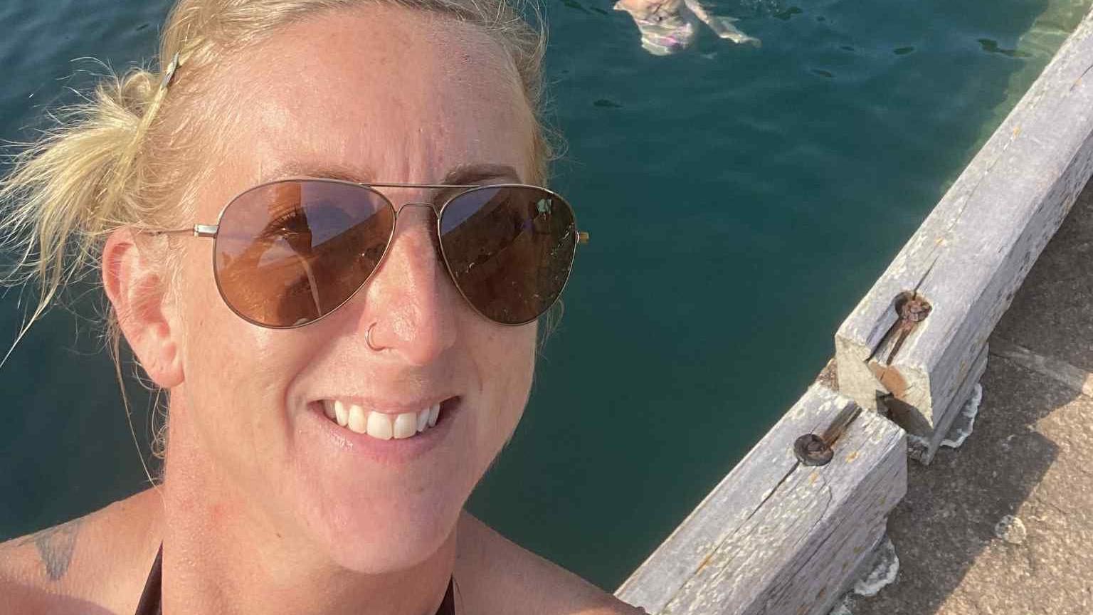 Helen Le Poidevin, a lady with blonde hair and sunglasses looking at the camera. She's standing in front of three people swimming in Sark's Creux Harbour, where there are five small boats floating.