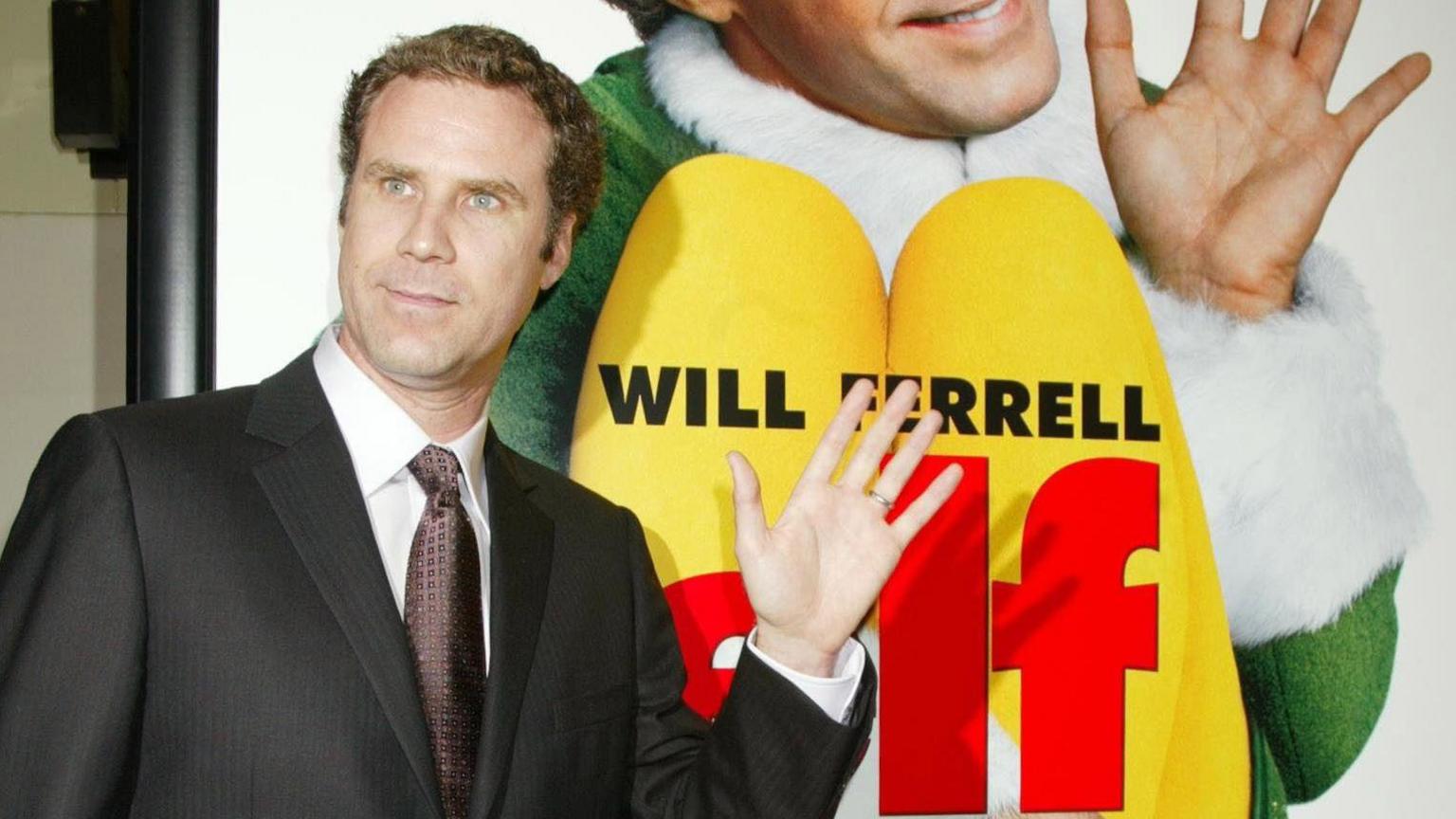 Actor Will Ferrell smiles and waves next to a poster of himself in the film promotion for Elf